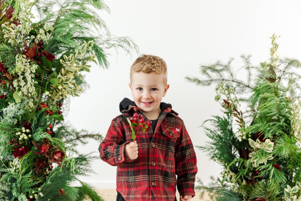 Child photographed in front of a holiday greenery backdrop with soft botanical textures.