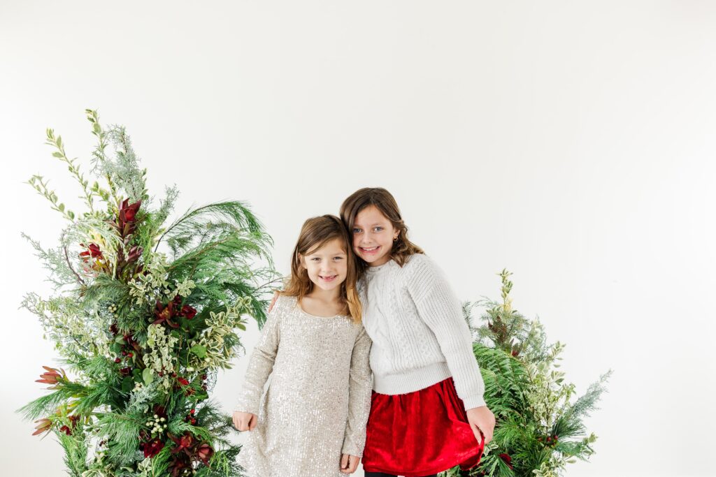 Smiling children in front of organic festive greenery for holiday portraits.