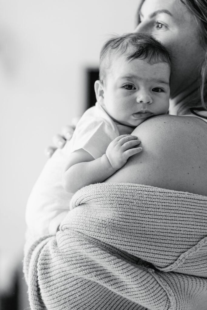 Mother holding her newborn in soft natural window light during an in‑home session.